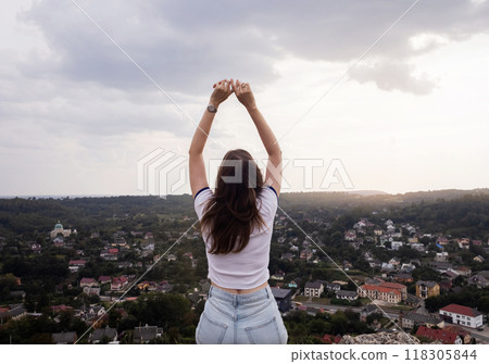 Young woman sits on a mountain with her hands raised like a winner. A gesture of freedom and satisfaction from the seen landscape. A beautiful view of the city from above Young woman sits on a mountain with her hands raised like a winner. A gesture of freedom and satisfaction from the seen landscape. A beautiful view of the city from above 118305844