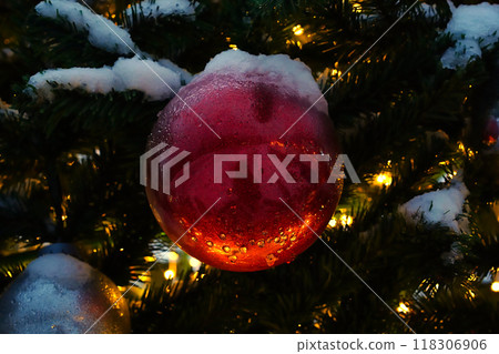 Christmas tree bauble red ball covered with snow and ice, lit with garlands in the evening. Horizontal photo template for New Year or Christmas card. Artificial, decorated green plastic Xmas tree. Christmas tree bauble red ball covered with snow and ice, lit with garlands in the evening. Horizontal photo template for New Year or Christmas card. Artificial, decorated green plastic Xmas tree. 118306906