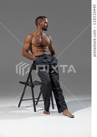 Man leans casually against chair, hands in pockets, showcasing his athletic build. Shirtless guy dressed pants. 118310440