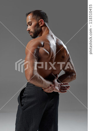 Rear view photo of young man with muscular body posing shirtless holding hands behind back against grey studio background. Rear view photo of young man with muscular body posing shirtless holding hands behind back against grey studio background. 118310455