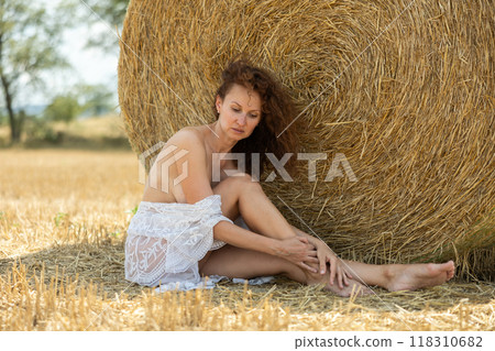Half-naked girl in white lacy negligee poses sitting in nature in field Half-naked girl in white lacy negligee poses sitting in nature in field 118310682