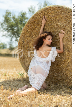 Back view of woman in white peignoir kneeling at haystack 118310724
