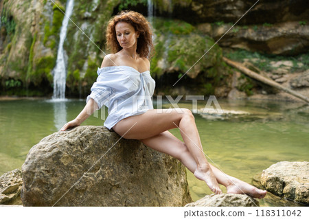 Half-naked girl in light linen shirt poses sitting in nature against backdrop of waterfall Half-naked girl in light linen shirt poses sitting in nature against backdrop of waterfall 118311042