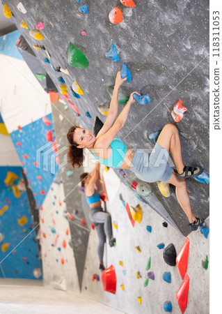 Adult woman is mastering climbing on training wall in gym, side view. Young woman holds on tightly to ledges and strives for top of bouldering route 118311053