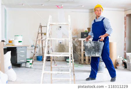 Worker in blue overalls and hardhat dragging various painting tools in room being renovated Worker in blue overalls and hardhat dragging various painting tools in room being renovated 118311229