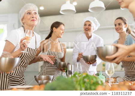 Aged woman holding wine and talking to other cooking course members whipping souce in bowls Aged woman holding wine and talking to other cooking course members whipping souce in bowls 118311237