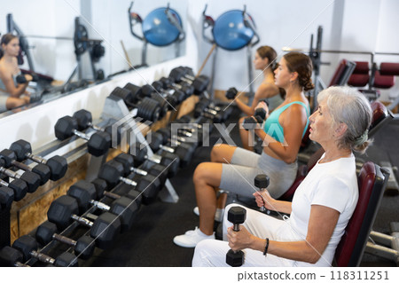 Slender senior woman sit and trains muscles of arms and back using dumb-bell shaped figure weighting Slender senior woman sit and trains muscles of arms and back using dumb-bell shaped figure weighting 118311251