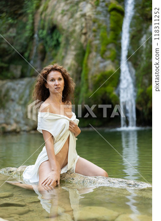 Half-naked girl in light dressing gown poses sitting in nature against backdrop of waterfall 118311252