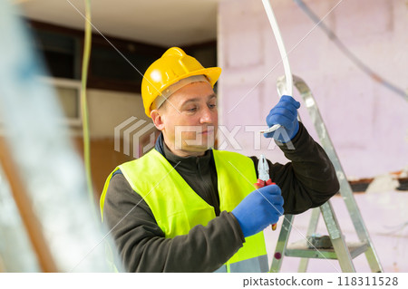 Electrician in protective uniform and helmet with pliers in his hands prepares electrical wiring in house under construction 118311528