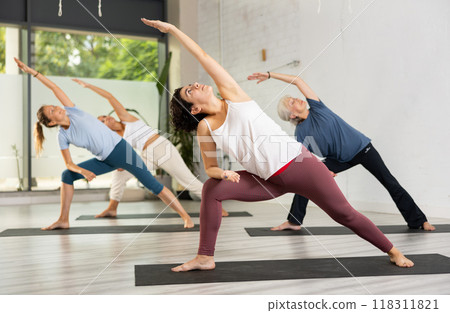 Young Latin female practicing Revolved Side Angle Pose Parivrtta Parsvakonasana, balancing posture of Hatha yoga during group workout in studio Young Latin female practicing Revolved Side Angle Pose Parivrtta Parsvakonasana, balancing posture of Hatha yoga during group workout in studio 118311821