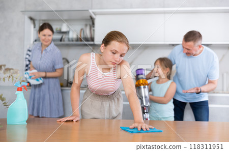 Bored teenage girl wiping table while cleaning apartment with family Bored teenage girl wiping table while cleaning apartment with family 118311951