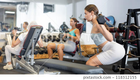 Slender girl trains muscles of arms and back using barbell and differentiated weighting in gym 118312155