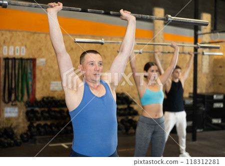 Motivated man practicing exercises with rod near other people in gym Motivated man practicing exercises with rod near other people in gym 118312281