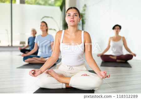 Calm young woman sitting in Lotus Pose Padmasana on mat during meditation and group yoga class in fitness studio 118312289