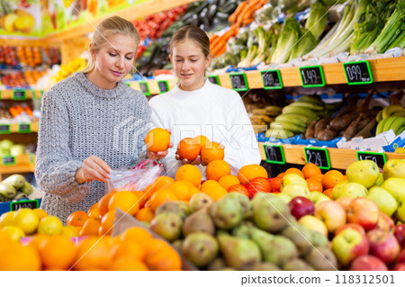 Cheerful teen girl with mother choosing sweet oranges in greengrocery 118312501