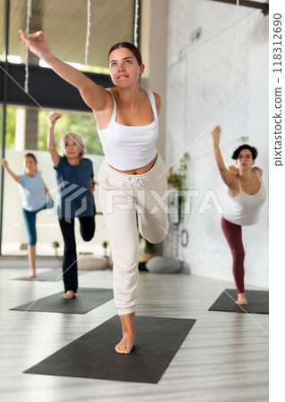 Young woman balancing in Natarajasana (Dancer Pose or Lord of the Dance Pose) during group yoga class in fitness studio 118312690