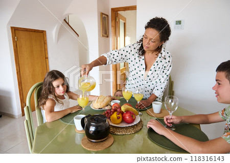 A mother serves orange juice to her children at a breakfast table with fruits and bread, creating a warm family atmosphere. 118316143