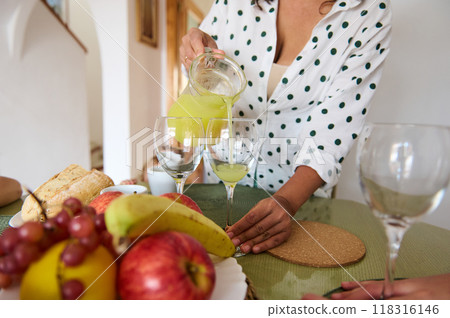 A woman pours fresh juice into glasses on a table adorned with fruit and bread, creating a serene and inviting kitchen scene. A woman pours fresh juice into glasses on a table adorned with fruit and bread, creating a serene and inviting kitchen scene. 118316146
