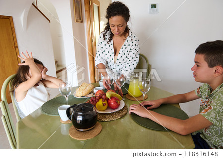A mother prepares breakfast for her children at a dining table, featuring fresh fruit, bread, and juice. The image captures a cozy family moment. 118316148