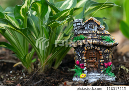 Miniature statuette of a fairy gnome house stands under the hosta plant on the brown mulch and decorates the flower bed. Miniature statuette of a fairy gnome house stands under the hosta plant on the brown mulch and decorates the flower bed. 118317215