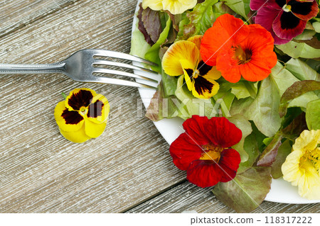 Table top view of the plate with salad made of edible flowers (nasturtium, pansy) and lettuce on the wooden table with a fork. Table top view of the plate with salad made of edible flowers (nasturtium, pansy) and lettuce on the wooden table with a fork. 118317222