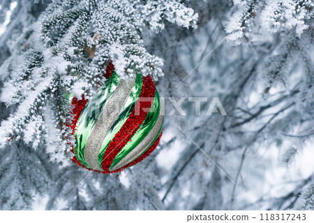 Large Christmas ball of geometric pattern with yellow, red, green stripes is hanging on the spruce tree branch covered with snow. 118317243