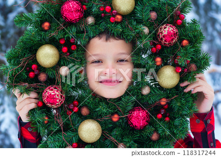 A boy in red plaid shirt is outside near snowy tree holding a handmade Christmas wreath made of pine branches and red and golden baubles. 118317244