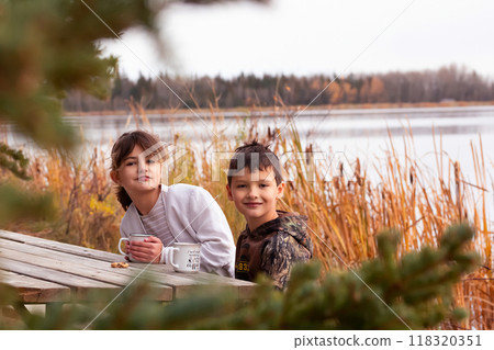 Two kids with mugs are at the table at the park near the lake in fall. Two kids with mugs are at the table at the park near the lake in fall. 118320351