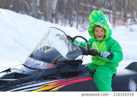 Ten year old boy is sitting on a black snowmobile in a cool bright green dragon-like snowsuit  and laughing in the winter forest. 118320359