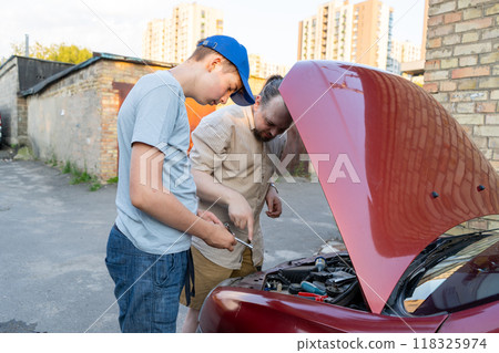 Dad and teenage son work together on the street near the garage. Outdoor activity. Family spend time. Helping and teach. The car hood is open. Engine repair and oil change. 118325974