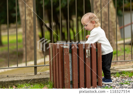 Adorable caucasian child looking into a trashcan with curiosity on a spring day. Cute blonde kid exploring the world around him in a park. Beautiful boy searching through a trash can in a garden. 118327554
