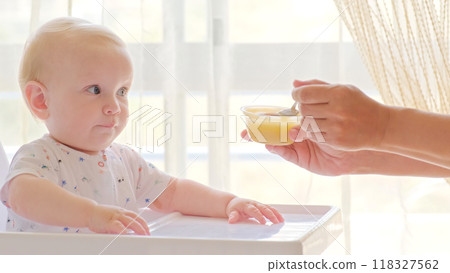 Cute baby is sitting on a high chair waiting for a meal. Beautiful white-haired toddler is sitting in a high children's chair at home. 118327562
