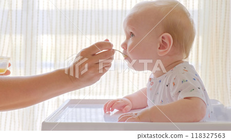 Mother is Feeding a baby from a spoon. Mom feeds a 9 month baby with fruit puree from a spoon, close-up, high key. Infant boy eats sitting on baby's chair. Mother cares about little son. 118327563