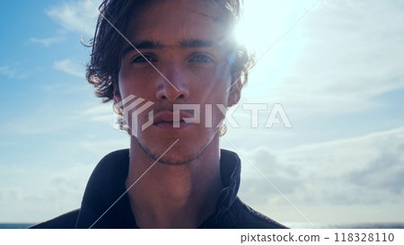 Young man with blown brown hair against the blue sky on summer sunny day. Stylish man in black jacket looks at the camera, low angle shot. Handsome guy with long hair poses outdoors. Young man with blown brown hair against the blue sky on summer sunny day. Stylish man in black jacket looks at the camera, low angle shot. Handsome guy with long hair poses outdoors. 118328110