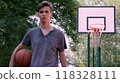 Teenager holds a basketball in his hand is on the sport ground and looking to the camera. Handsome Young man on a basketball ground with a basketball in his hands. Sport and activity concept. 118328111