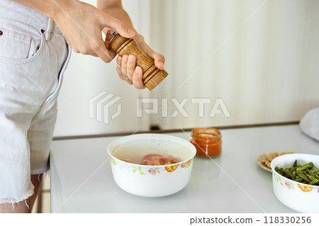 Person seasoning marinated chicken in a white bowl at home kitchen with various condiments nearby 118330256