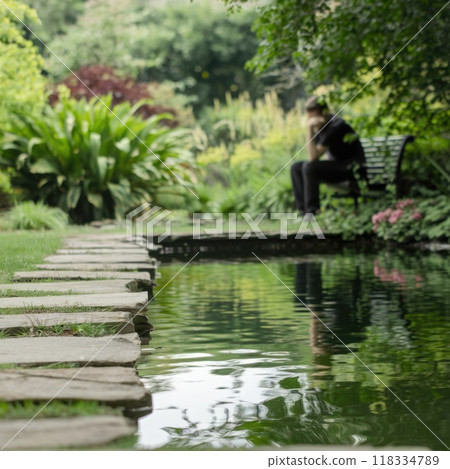 A peaceful garden with a stone pathway leading to a still pond, reflecting the sky and greenery. A person sits on a bench, lost in quiet contemplation. 118334789