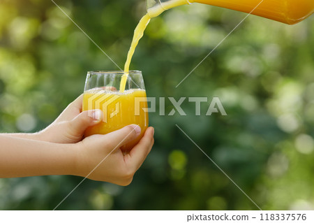 child hands holding glass and orange juice pouring from jug with green garden on the background child hands holding glass and orange juice pouring from jug with green garden on the background 118337576