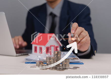 Businessman in suit analyzing increasing real estate market trends using a model house and stacked coins with an upward arrow and percentage symbols 118338937