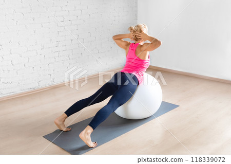 Fit adult woman in pink tank top practice back extension exercise with hands behind her head using fitness ball in loft studio indoor, back view. Fit adult woman in pink tank top practice back extension exercise with hands behind her head using fitness ball in loft studio indoor, back view. 118339072