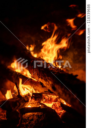 Flickering flames illuminate wooden logs, providing warmth and a cozy atmosphere on a cool evening outdoors. Flickering flames illuminate wooden logs, providing warmth and a cozy atmosphere on a cool evening outdoors. 118339686