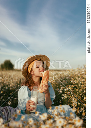Girl in blue linen dress and canotier straw hat sits in daisy field and eats baguette with milk, vintage photo Girl in blue linen dress and canotier straw hat sits in daisy field and eats baguette with milk, vintage photo 118339934