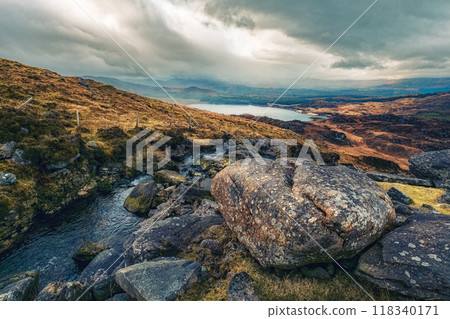A breathtaking view of rocky terrain leads down to a serene lake, with rolling hills in the background under a moody sky filled with clouds. 118340171