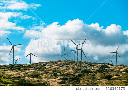 Wind turbines are seen on a rugged hillside, with a partly cloudy sky above. Wind turbines are seen on a rugged hillside, with a partly cloudy sky above. 118340175