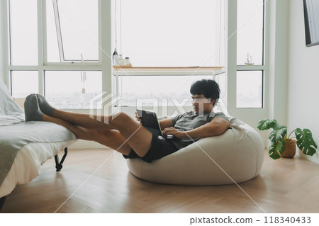 Man working with his laptop lying on the bean bag sofa in living room. 118340433