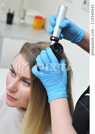 A dermatologist holds a dermatoscope in his hands and examines the structure of a young woman's hair. Trichoscopy is the science of hair structure. 118340842
