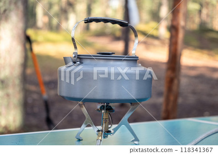 A silver kettle sits on a camping stove surrounded by trees, ready for morning coffee while the sun rises in the background. A silver kettle sits on a camping stove surrounded by trees, ready for morning coffee while the sun rises in the background. 118341367