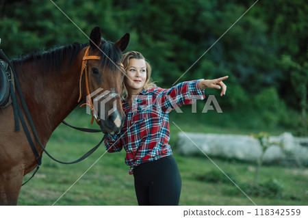 Happy blonde with horse in forest. Woman and a horse walking through the field during the day. Dressed in a plaid shirt and black leggings. Happy blonde with horse in forest. Woman and a horse walking through the field during the day. Dressed in a plaid shirt and black leggings. 118342559