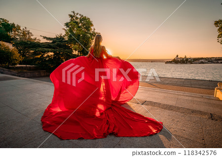 Sunrise red dress. A woman in a long red dress against the backdrop of sunrise, bright golden light of the sun's rays. The concept of femininity, harmony. 118342576