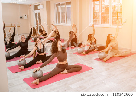 A group of six athletic women doing pilates or yoga on pink mats in front of a window in a beige loft studio interior. Teamwork, good mood and healthy lifestyle concept. 118342600